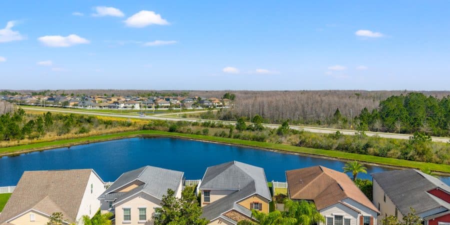 Bright suburban neighborhood with colorful houses, a serene pond, and lush greenery under a clear blue sky.